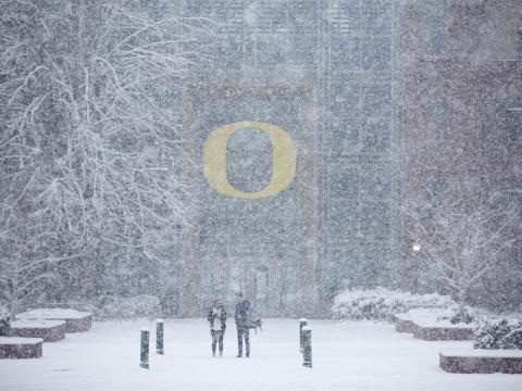 Two people stand in the falling snow outside of the Lillis building.